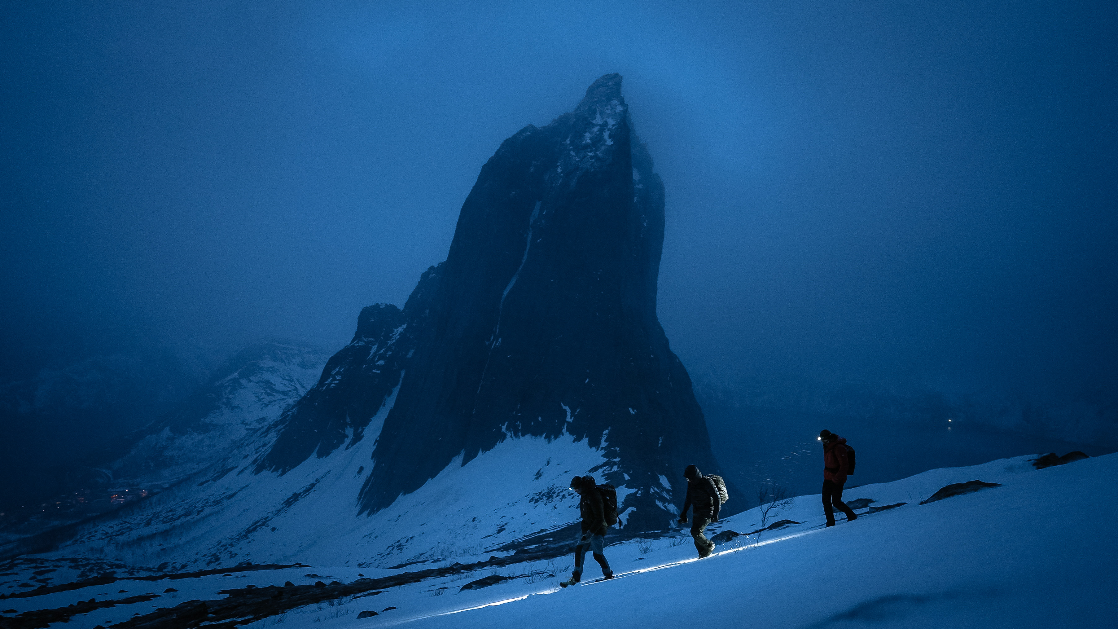Three people walk down a steep, snowy slope at dusk